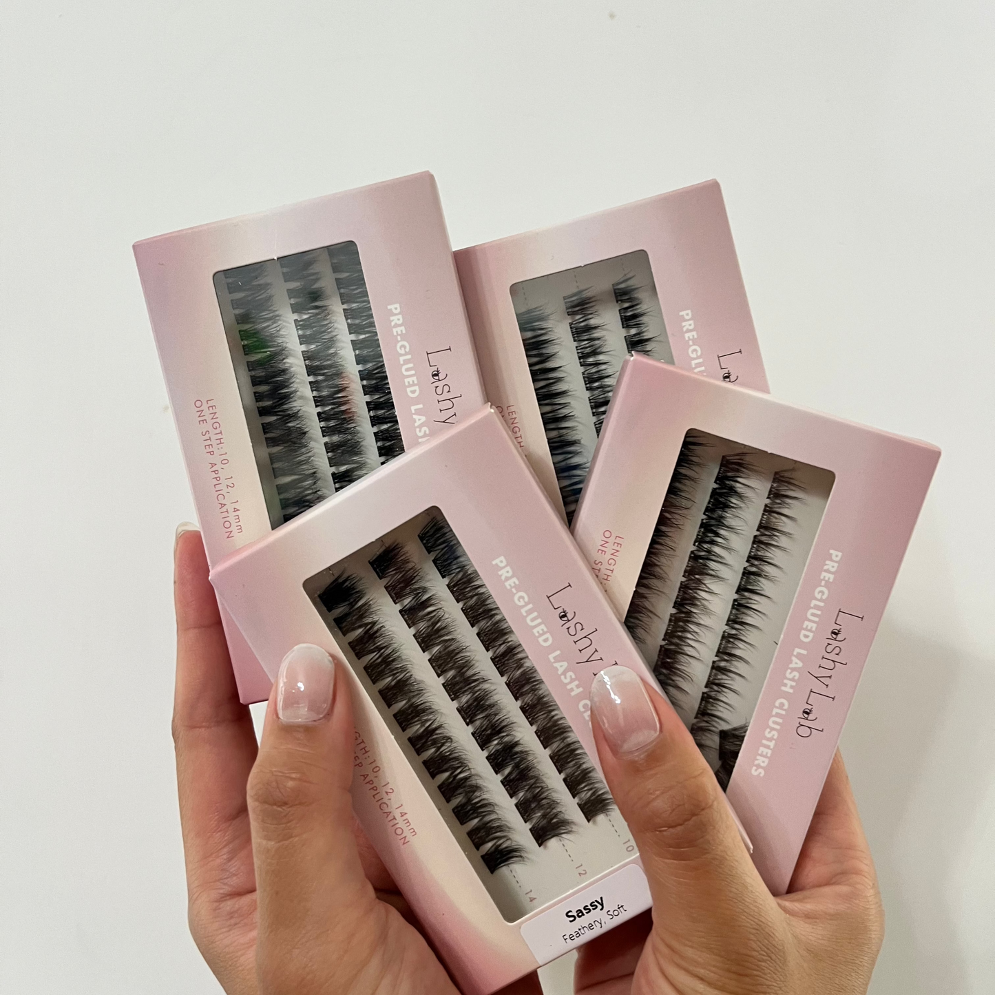 A person with manicured nails holds four Lashy Lab PRE-GLUED LASHES MIX-MATCH BUNDLE boxes, each displaying lash clusters through clear windows—perfect for easy DIY eyelash extensions.