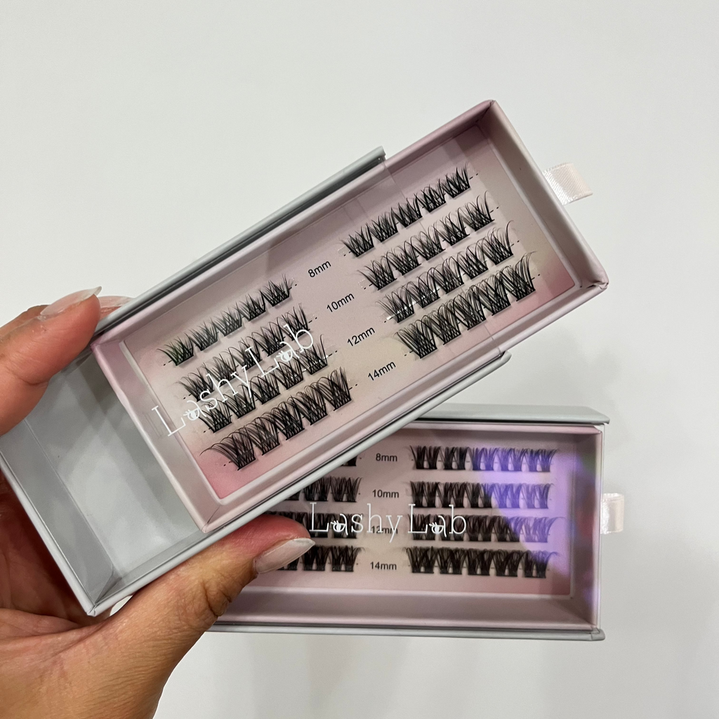 A hand holds two open PRO CLUSTER LASHES STARTER BUNDLE boxes by Lashy Lab, each featuring DIY eyelash extensions in 8mm–14mm lengths on a pink base. The white boxes with ribbon tabs offer lashes that last for weeks.