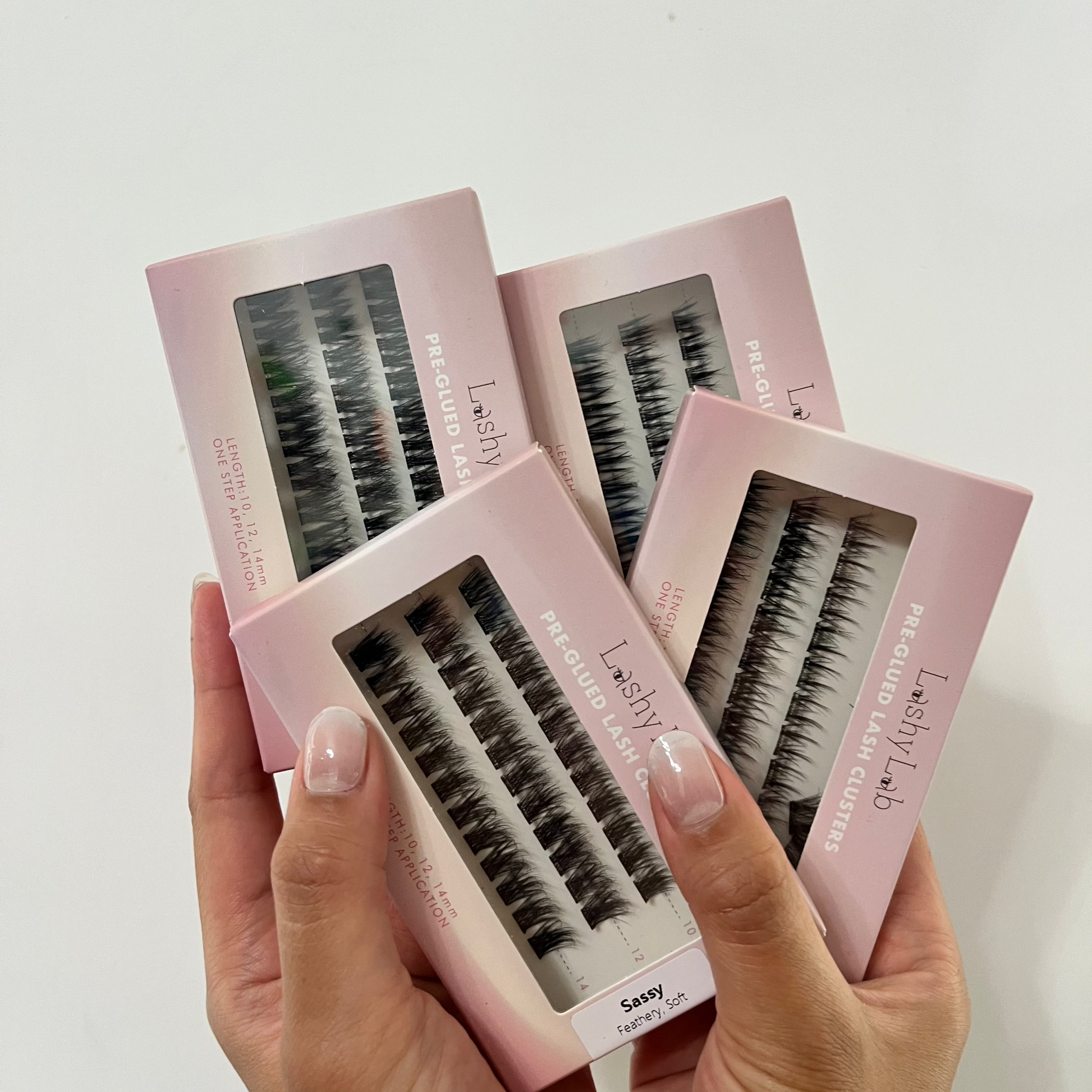 A person with manicured nails holds four Lashy Lab PRE-GLUED LASHES MIX-MATCH BUNDLE boxes, each displaying lash clusters through clear windows—perfect for easy DIY eyelash extensions.