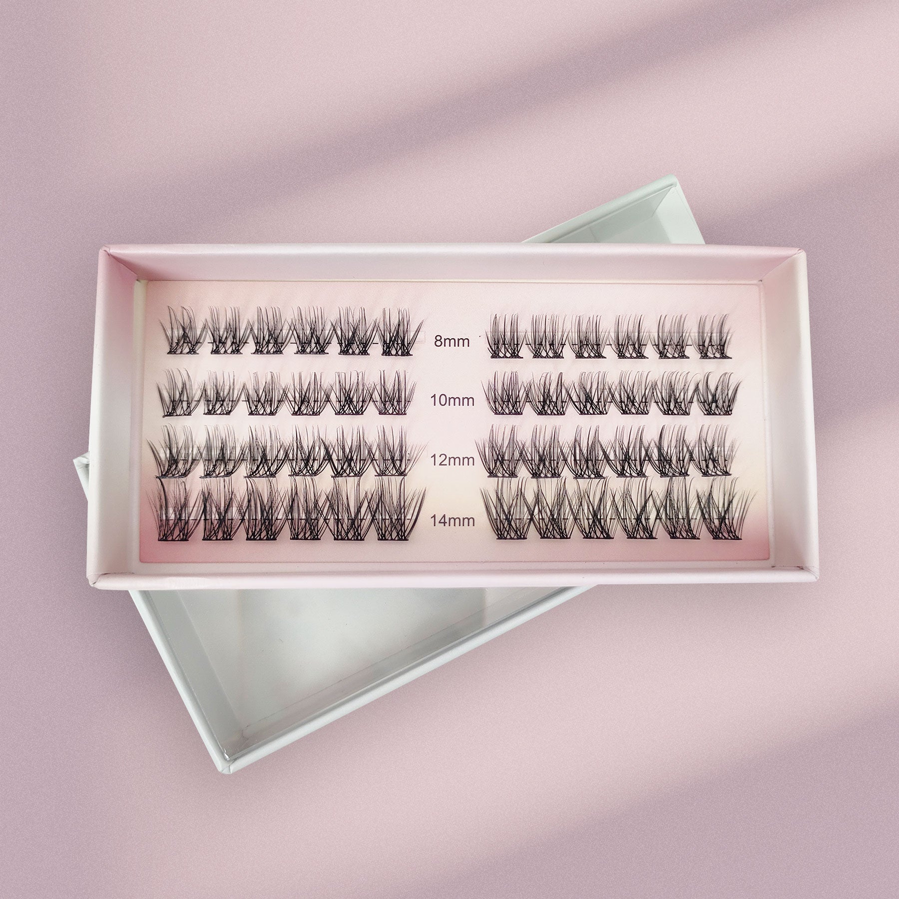 An open box of Lashy Labs CHIC GIRL Wispy, Trendy, Cat Eye lash extensions rests on a pink surface, showcasing four rows of DIY lashes in 8mm, 10mm, 12mm, and 14mm lengths that stay on for weeks.