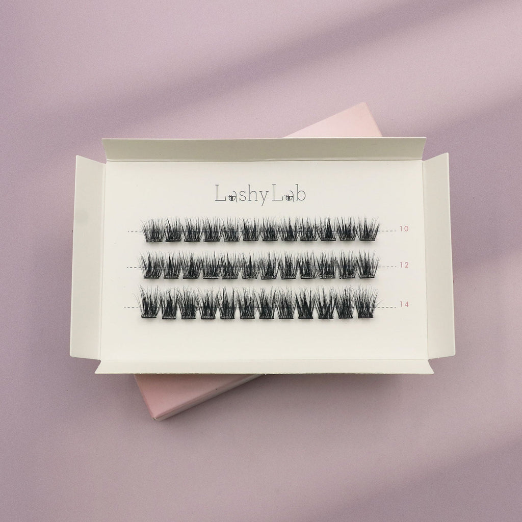 A white box labeled Lashy Lab SASSY PRE-GLUED LASHES SET holds three rows of pre-glued cluster lashes in assorted lengths, displayed on a pastel lilac and pink background for easy application.
